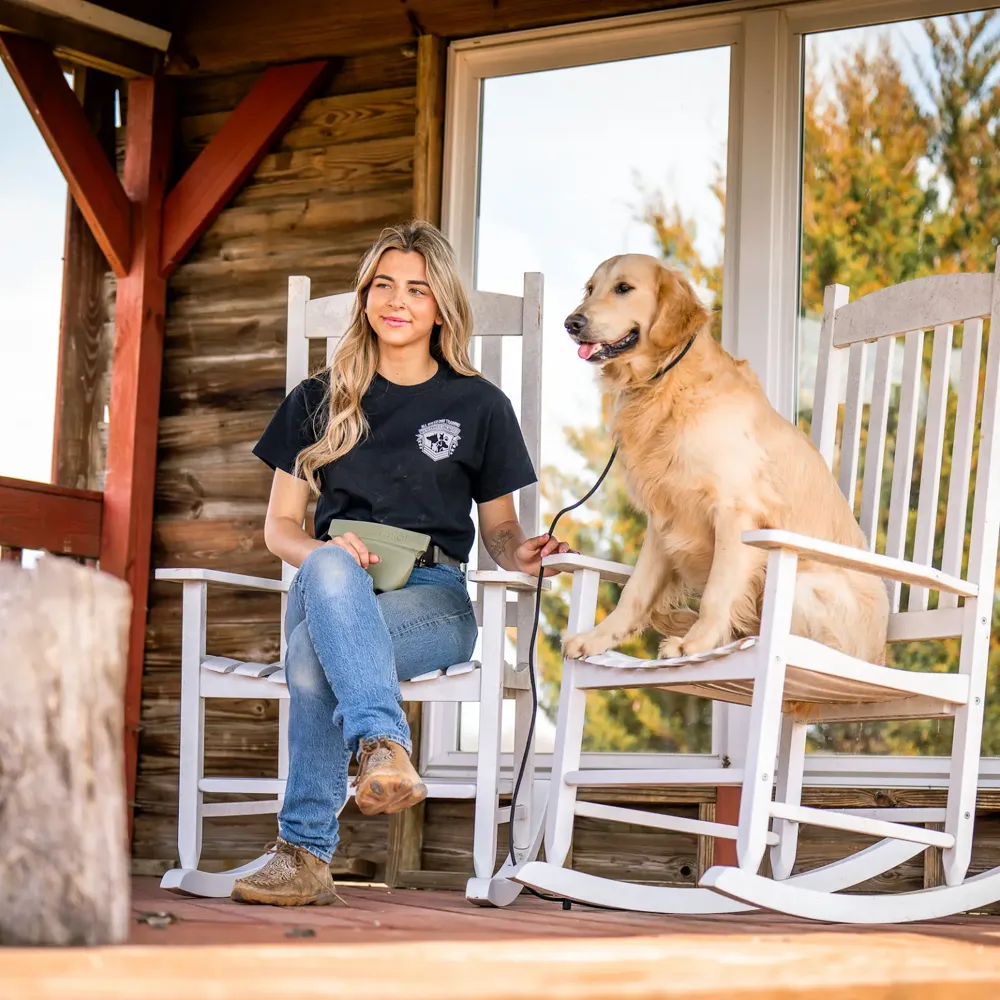 A trainer sitting in a white rocking chair on a log-cabin porch with a golden retriever resting beside her