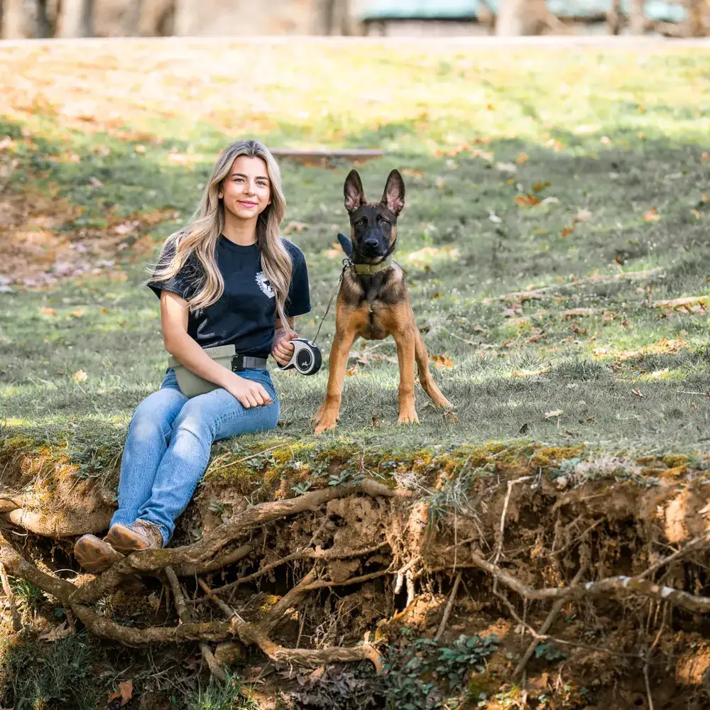 A trainer sitting on a grassy hillside with a young German shepherd standing beside her